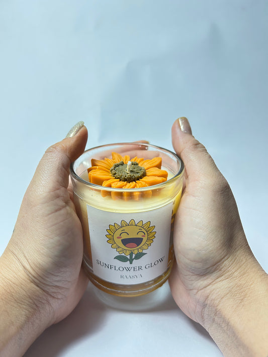 Candle with sunflower design held by two hands against a light background